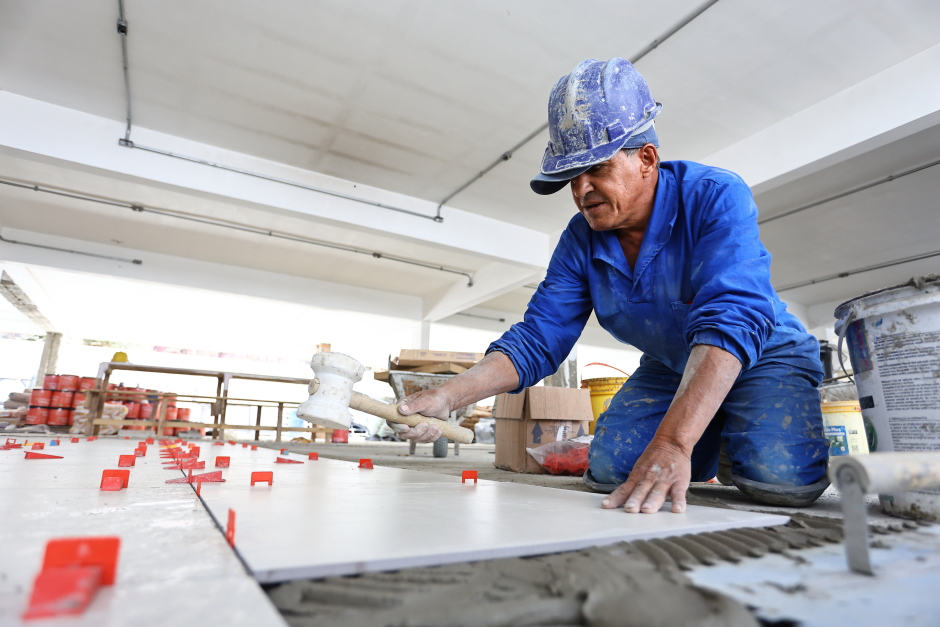 Pedreiro vestido com uniforme azul assenta piso com martelo de borracha em obra de uma escola municipal