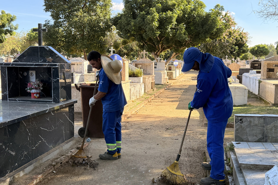 Dois funcionários da Urbanizadora Municipal aparecem vestidos com o uniforme azul varrendo resíduos no cemitério Colônia Paraíso