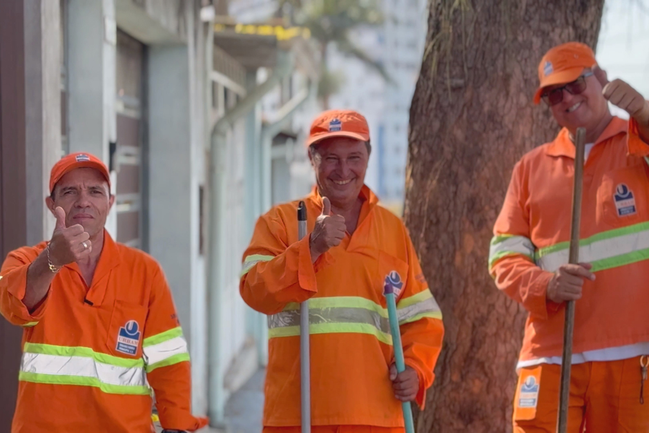 Três auxiliares de serviços gerais, uniformizados em seus trajes laranja da Urbam, posam para a imagem enquanto trabalham na varrição